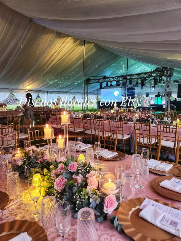 Montaje de mesas con flores rosas y velas frente a un escenario iluminado.
