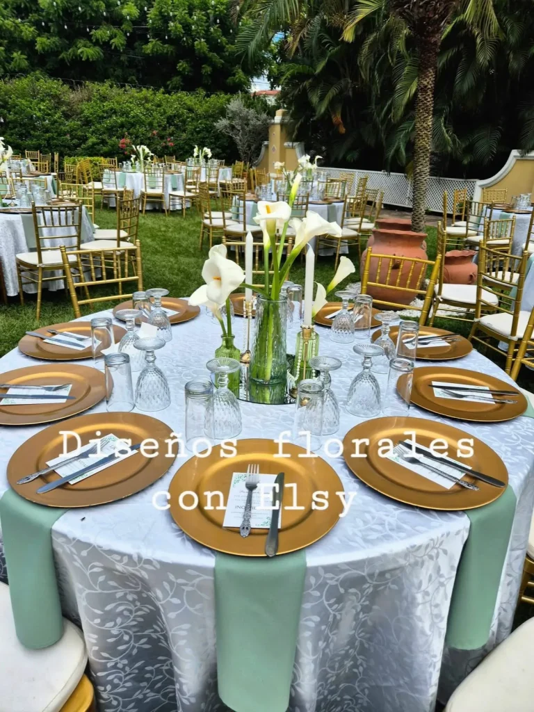 Mesa redonda en el jardín con alcatraces blancos, cristalería y platos dorados.