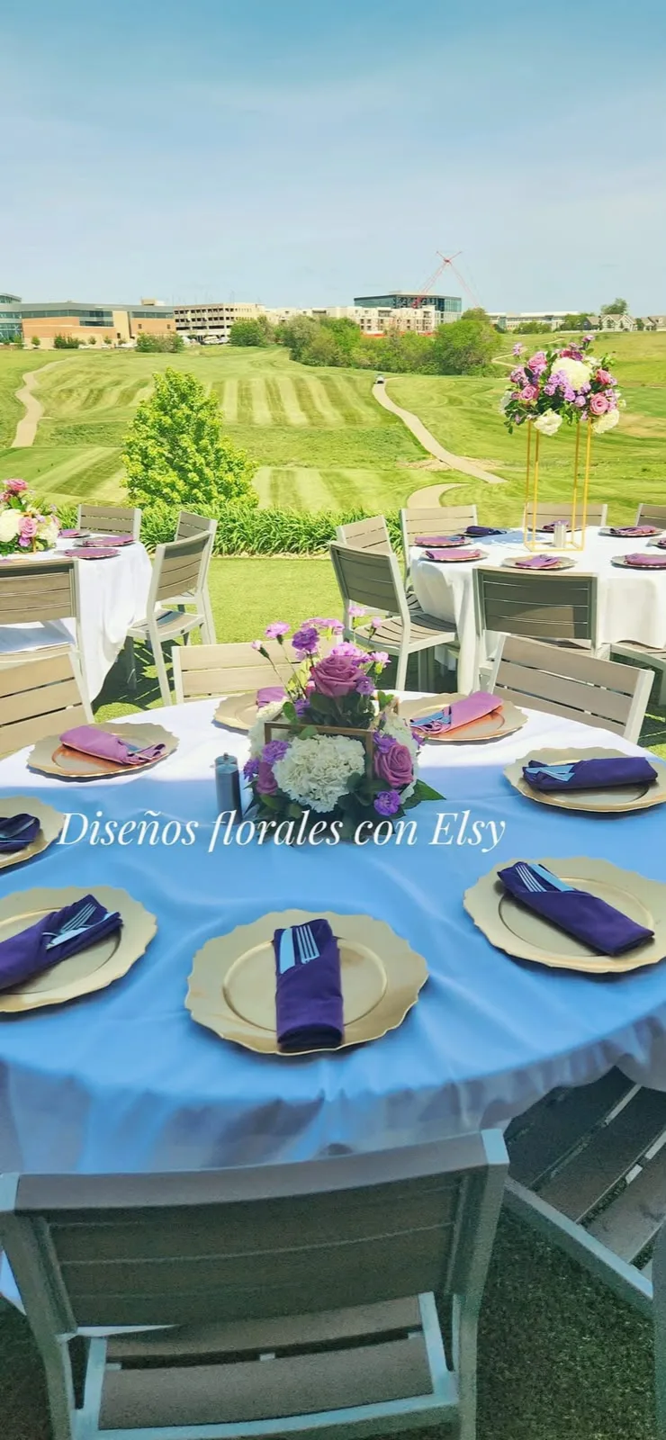 Mesas redondas al aire libre con manteles blancos, servilletas lilas y centros florales en bases doradas.
