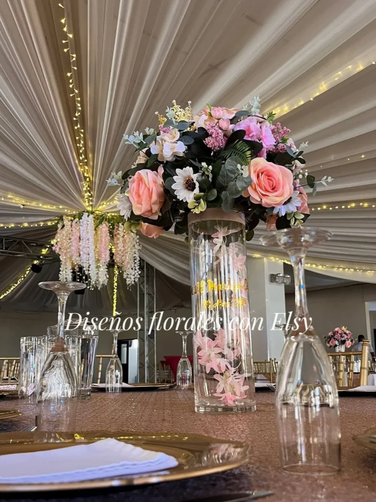Centro floral alto en tonos rosa y blanco sobre un cilindro de vidrio, bajo techo decorado con telas y luces.