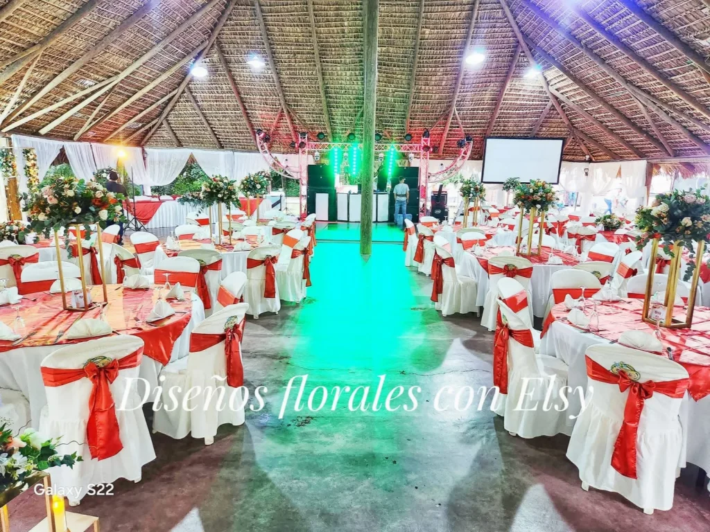 Mesas con manteles rojos y sillas blancas con lazos rojos bajo techo de palma, con flores y luces al fondo.