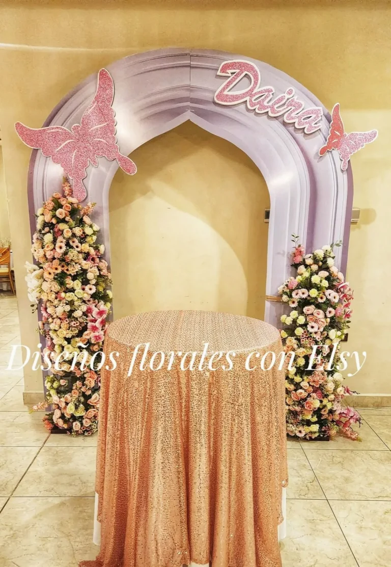 Arco en tonos rosa con mariposas y arreglos florales a los lados de una mesa cubierta con tela brillante.