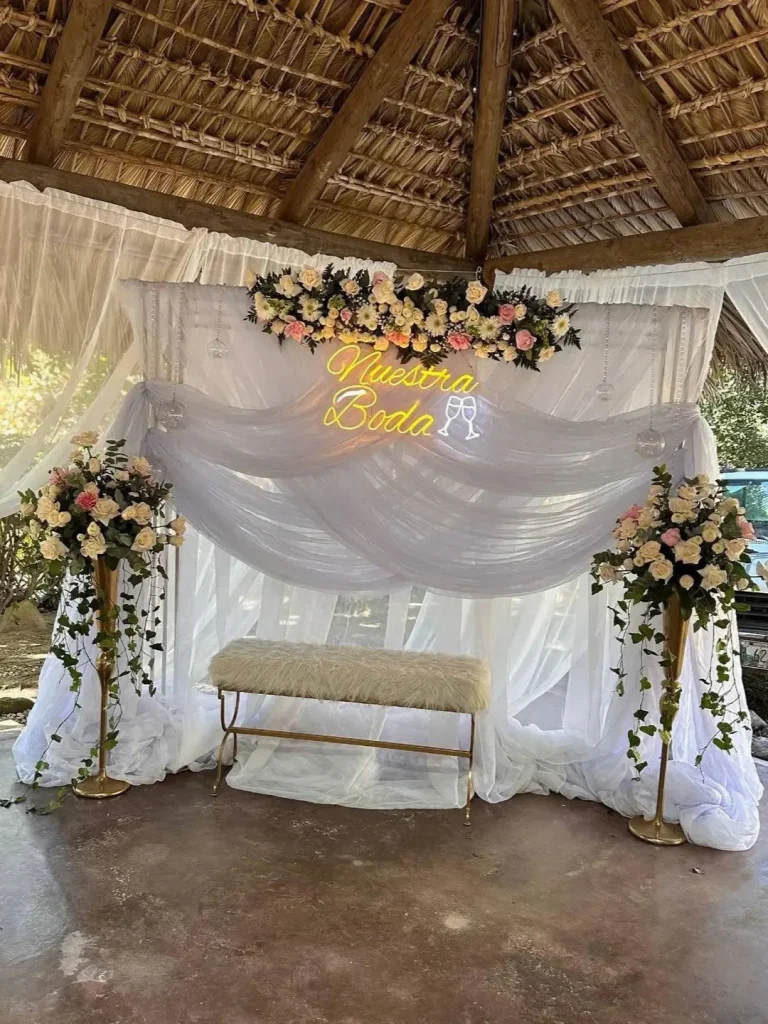 Fondo de boda con telas blancas, flores claras, banco y letrero luminoso Nuestra Boda bajo cubierta de palma.